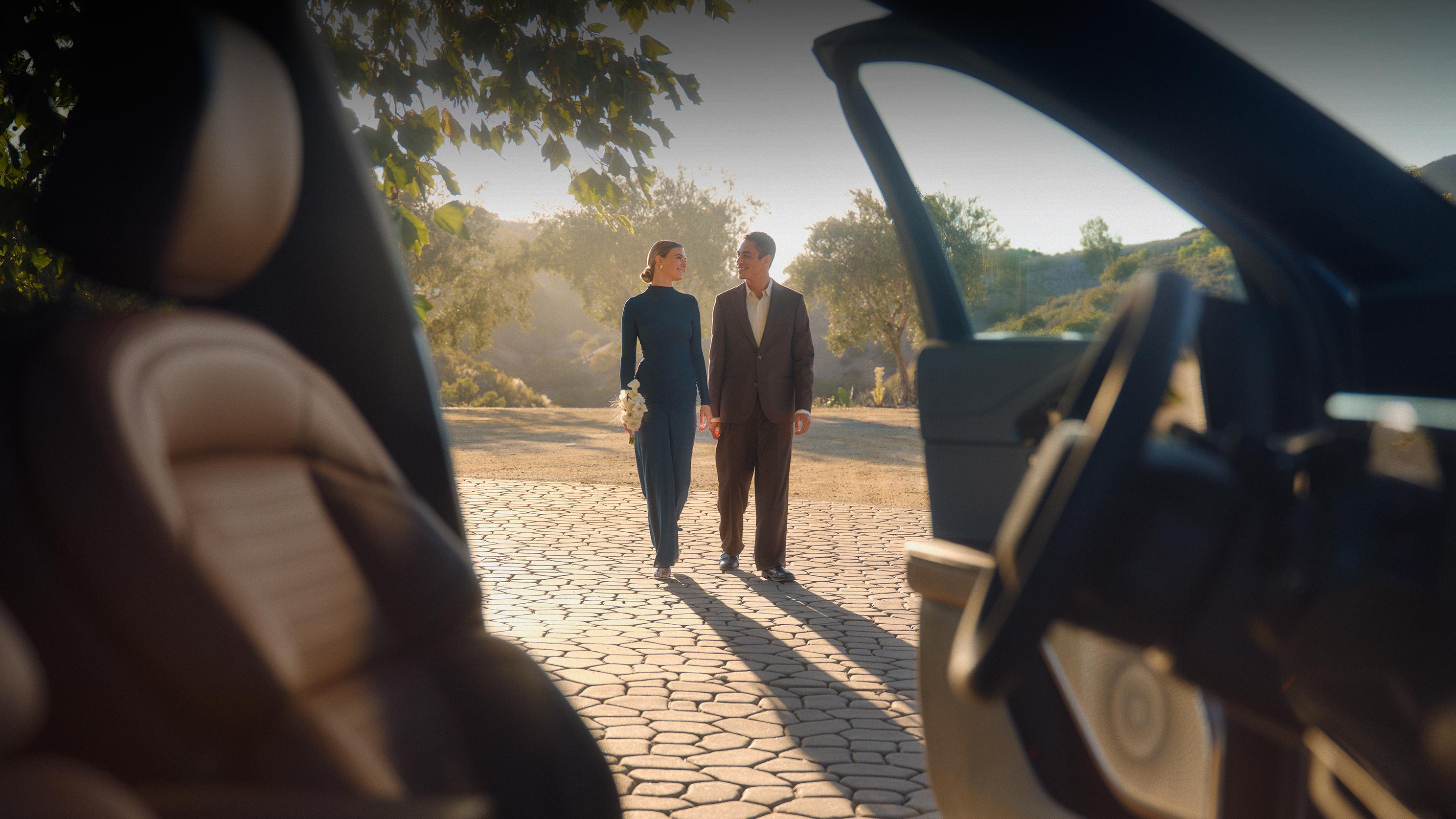 Un homme et une femme se tenant la main et se souriant en marchant en plein air, vus depuis l’intérieur d’une voiture.