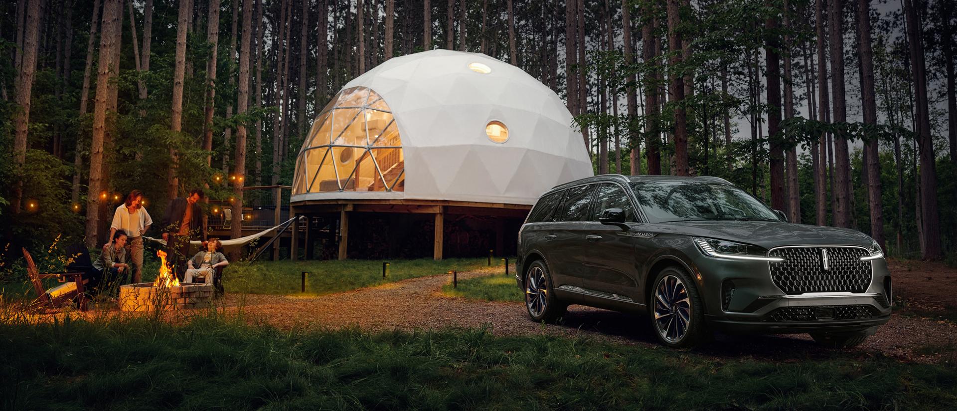 Un Lincoln Aviator® 2027 stationné à côté d’un site de camping luxueux pendant qu’une famille profite ensemble d’un feu de camp