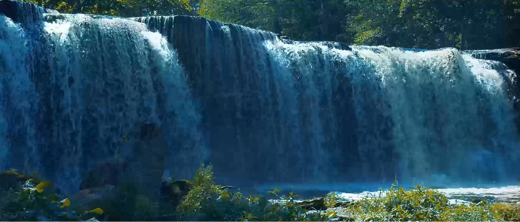Vignette vidéo de la thématique élargie des chutes d’eau montrant une chute d’eau en cascade qui coule sur une falaise rocheuse dans la verdure ci-dessous.