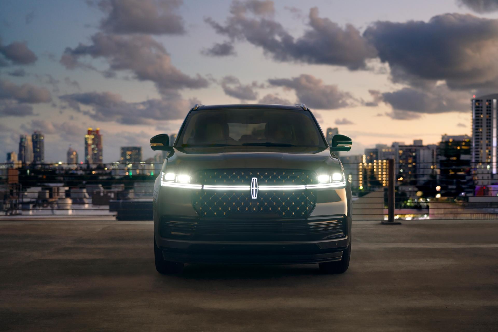 The front of a 2026 Lincoln Navigator® has it's headlamps and Lincoln Star illuminated against a city backdrop at dusk